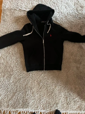 Svart hoodie från Polo Ralph Lauren -  Säljer nu en fin svart hoodie från Polo Ralph Lauren med dragkedja, två fickor framtill och vit snörning i huvan. Materialet är mjukt och bekvämt, perfekt för chill dagar. Röd Ralph lauren logga. Nästan aldrig använd väldigt fint skick 