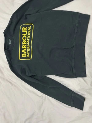 Svart sweatshirt från Barbour - Cool svart sweatshirt från Barbour International med stor gul logga framtill. Tröjan har rund halsringning, långa ärmar och ribbade muddar vid ärmslut och nederkant. Perfekt för en avslappnad streetwear-look.