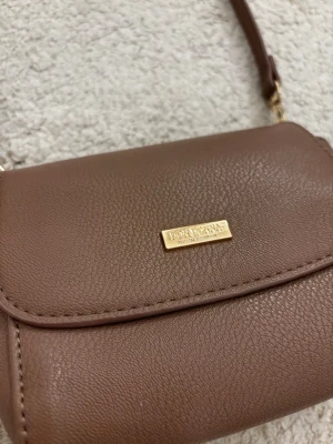 Brun/beige axelväska från Don Donna - Snygg brun/beige axelväska från Don Donna i ett stilrent och kompakt format. Väskan är gjord i strukturerat konstläder med justerbar axelrem och guldfärgade metalldetaljer. Perfekt för dig som gillar minimalistisk design och vill ha en väska som funkar till det mesta.