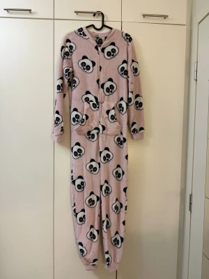 Rosa panda-mönstrad onesie - Mysig rosa onesie med svartvita pandaansikten över hela plagget. Långärmad och helkropp, med dragkedja framtill och två fickor. Perfekt för att chilla hemma och hålla sig varm. Materialet är mjukt fleece som känns skönt mot huden.