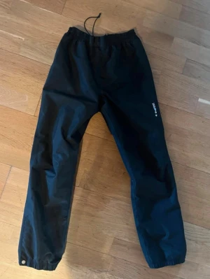Svarta tävkbyxor från Everest - Säljer ett par svarta vindbyxor från Everest med resår i midjan och benslut samt dragsko upptill. Byxorna är tillverkade i ett lätt och vindtätt material, perfekta för blåsiga dagar. (Aldrig använda) Kom gärna med prisförslag eller frågor.