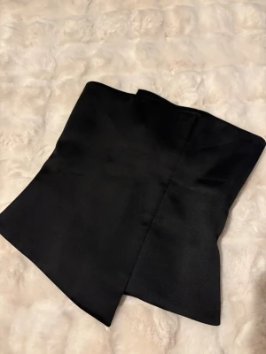 Svart asymmetrisk tubtopp - Snygg svart tubtopp med asymmetrisk nederkant i ett glansigt tyg. Toppen är axelbandslös och har en stilren, minimalistisk design som passar perfekt till trendiga outfits. Den är i storlek 38 men skulle säga att den är liten i storlek och därför mer är som 34/36