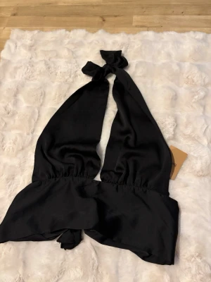 Svart omlott linnetopp med knyt - Trendig svart linnetopp med knyt både i nacken och i ryggen. Toppen har djup v-ringning och är ärmlös, perfekt för en snygg och stilren look. Tillverkad i ett glansigt material som ger en lyxig känsla. Storleken är 38, men går att justeras vid knytningarna