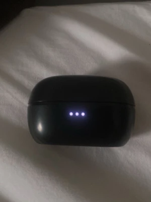 Svart trådlöst hörlursfodral - Säljer ett svarta jbl, ovalt fodral till trådlösa in-ear hörlurar med LED-indikator på framsidan och USB-C laddningsport på baksidan. Fodralet är gjort av plast och har en stilren, minimalistisk design. Perfekt för dig som vill ha smidiga hörlurar till mobilen.