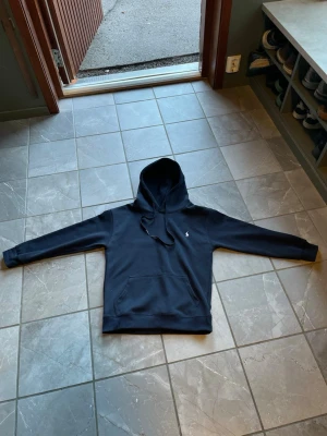 Mörkblå hoodie från Polo Ralph Lauren - Snygg mörkblå hoodie från Polo Ralph Lauren med klassisk huva och dragsnören. Tröjan har en liten broderad logotyp på bröstet och en stor magficka framtill. Materialet är mjukt och bekvämt, perfekt för chill dagar. Priset är inte hugget i sten vid snabb affär. 