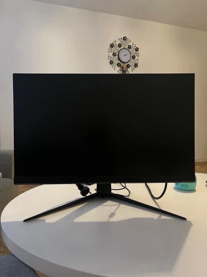 Svart datorskärm med stativ - Stilren svart datorskärm med tunn ram och bred fot i metall. Skärmen har en modern, minimalistisk design och passar perfekt för gaming eller arbete. Anslutningskablar syns på baksidan. Märke och exakt storlek framgår ej.