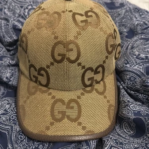 Beige och brun Gucci keps M - Säljer en snygg beige och brun keps från Gucci i storlek M. Kepsen har det klassiska GG-mönstret över hela ytan och detaljer i brunt skinn längs kanten. Materialet är slitstark bomull och polyester, perfekt för dig som vill ha en lyxig streetstyle-look.