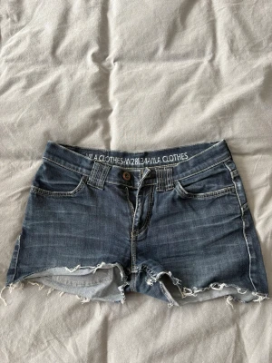 Low waisted shorts från Vila Clothes - Snygga low waisted jeansshorts från Vila Clothes med råa, fransiga kanter och klassisk femficksdesign. Shortsen har normal passform, bälteshällor och knappstängning. Perfekta för varma dagar och en avslappnad stil.