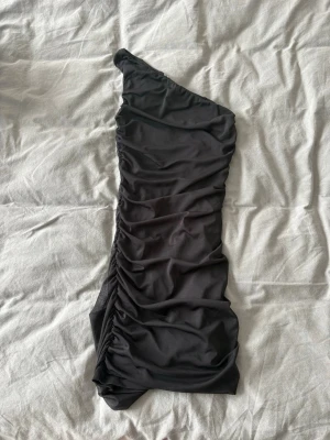 Svart one shoulder klänning - Snygg svart kortklänning med one shoulder-design och draperad effekt. Klänningen har en tight passform och är perfekt för dig som vill sticka ut. Tillverkad i ett mjukt och stretchigt material som sitter bekvämt.