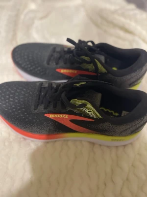 Brooks svarta sneakers med neonröd detalj - Snygga svarta sneakers från Brooks med neonröda och gula detaljer på sidan. Ovandelen är i mesh för bra andningsförmåga och sulan är vit med inslag av gult. Perfekta för dig som gillar sportig stil och vill sticka ut.