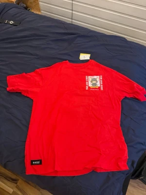 Röd Tom & Jerry t-shirt från BossH - Säljer en röd t-shirt med Tom & Jerry-tryck från BossH. Framsidan har en liten patch med Jerry och text, medan baksidan har ett stort tryck av Jerry i hoodie och texten 'Catch me if you can'. Perfekt för dig som gillar streetstyle och tecknade motiv.