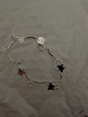 Silverarmband med nalleberlocker - Snyggt armband i silverfärg med små nalleformade berlocker och glittrande detaljer. Armbandet har en tunn kedja och justerbar längd med karbinlås. Perfekt accessoar för dig som gillar söta och lekfulla smycken.