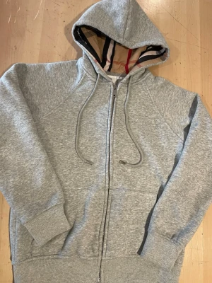 Grå zip-hoodie från Burberry - Snygg grå zip-hoodie från Burberry med klassiskt rutigt foder i huvan och dragsko. Storlek XS Tillverkad i mjuk bomullsblandning, har ribbade muddar och fickor framtill. Perfekt för en avslappnad och trendig look.
