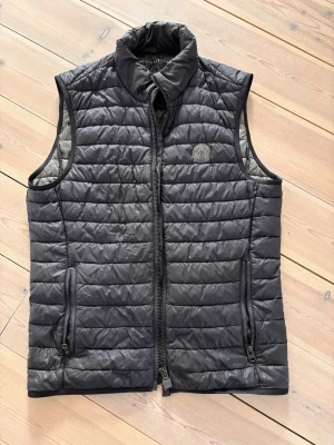 Dunväst från Marc o’polo - Snygg marinblå dunväst från Marc o’polo! Västen har två dragkedjeförsedda fickor framtill och en diskret logotyp på bröstet. Perfekt att slänga över en hoodie eller tröja för extra värme. Materialet är lätt och smidigt.