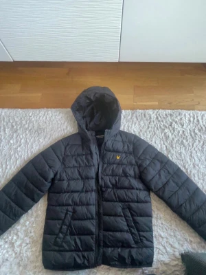 Svart dunjacka från Lyle & Scott - Snygg svart dunjacka från Lyle & Scott med huva och dragkedja framtill. Jackan har det klassiska gula logomärket på bröstet och quiltad design. Perfekt för kalla dagar och håller dig varm hela vintern men även våren.