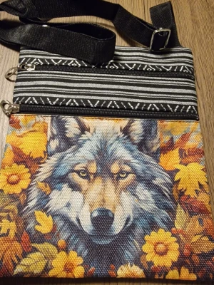 Axelväska med vargmotiv och mönster - Cool axelväska i tyg med svartvit etnisk mönstring och ett färgstarkt tryck av en varg bland gula blommor på framsidan. Väskan har justerbar svart axelrem och två dragkedjefack. Bredd 19 cm. Höjd 23,5 cm. Perfekt för dig som gillar unika accessoarer och djurmotiv.