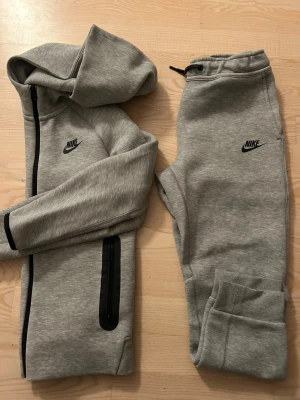 Grått set från Nike med huva - Snyggt grått set från Nike med zip-hoodie och matchande byxor. Hoodien har svart dragkedja och fickor, samt Nike-logga på bröstet. Byxorna har resår i midjan med snörning och Nike-logga på benet. Perfekt för en sportig och avslappnad stil.