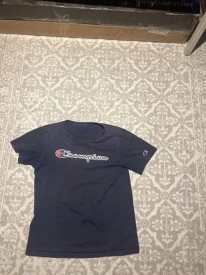 Marinblå Champion t-shirt - Snygg marinblå t-shirt från Champion med det klassiska logotrycket över bröstet och en liten logga på ärmen. T-shirten har rund hals och korta ärmar, perfekt för en avslappnad och sportig stil.