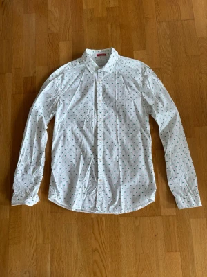 Vit skjorta från Scotch & Soda - Vit långärmad skjorta från Scotch & Soda med små gröna geometriska tryck. Klassisk krage och knäppning framtill. Skjortan är tillverkad i bomull och har en clean, modern look som passar till många stilar.