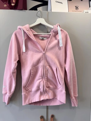 Ljusrosa hoodie med dragkedja - Mysig ljusrosa hoodie med dragkedja framtill och stora fickor. Tröjan har vit dragsko i huvan och ribbade muddar vid ärmar och nederkant. Insidan är mjuk och fleece-liknande för extra komfort. Perfekt för en avslappnad och chill stil.