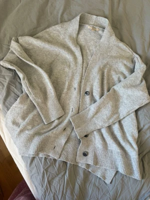 Ljusgrå kofta med knappar - Mysig ljusgrå kofta i mjukt material med djup V-ringning och stora ribbade muddar. Koftan har tre knappar framtill och en avslappnad passform, perfekt att slänga över en t-shirt. Enkel och stilren design som passar till det mesta.