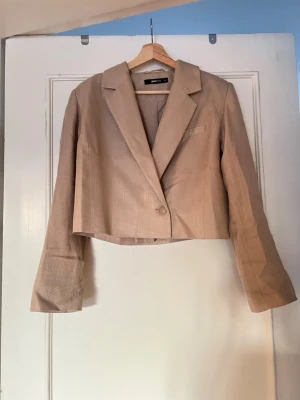 Beige cropped kavaj från Gina Tricot - Stilren beige cropped kavaj från Gina Tricot med klassisk krage och enkelknäppning. Kavajen har långa ärmar och en minimalistisk design som passar perfekt till trendiga outfits. Materialet är lätt och har en diskret struktur.