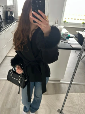 Svart kappa med fuskpälsdetaljer - Snygg svart kappa med stora fluffiga fuskpälsdetaljer vid ärmsluten och kragen. Kappan har knytband i midjan för en markerad siluett och en klassisk, elegant look. Perfekt för dig som vill ha en trendig och stilren ytterjacka till höst och vinter.