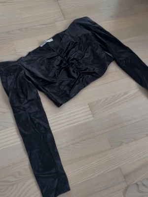 Svart långärmad satintopp - Snygg svart topp i satin med lång ärm och rynkade detaljer framtill. Toppen har en croppad passform och en lätt glansig yta som ger en lyxig känsla. Perfekt att styla med höga jeans eller kjol.