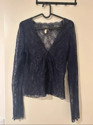 Mörkblå spetsblus med v-ringning - Snygg mörkblå långärmad topp i transparent spets med v-ringning och knappar framtill. Blusen har blommigt spetsmönster och är figurnära i modellen.