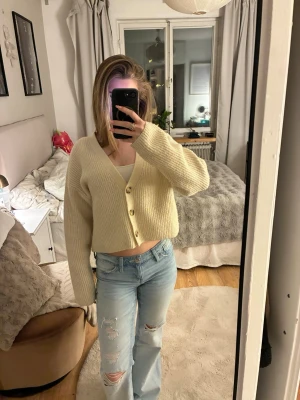 Krämvit cropped cardigan med knappar - Mysig krämvit cardigan i ribbstickat material med v-ringning och tre stora knappar framtill. Modellen är cropped och har långa, lite vidare ärmar för en avslappnad look. Perfekt att slänga över ett linne eller t-shirt.