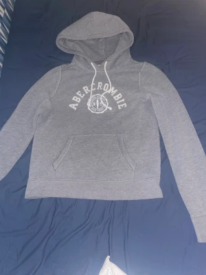 Abercrombie & Fitch Hoodie - Snygg grå hoodie från Abercrombie & Fitch med vit logga och text på bröstet. Klassisk känguruficka framtill och justerbar huva med vita snören. Perfekt för en avslappnad och trendig look. Mjuk bomullsblandning som känns skön mot huden. Strl S men passar xs också. Inga defekter.