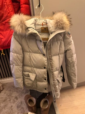 Ljusgrå dunjacka Parajumpers dam - Ljusgrå dunjacka från Parajumpers med stor fluffig pälskantad huva och quiltad design. Jackan har dragkedja, tryckknappar och två stora fickor framtill. Märkespatch på ärmen och detaljer i beige. Perfekt för kalla vinterdagar. Den är figursydd och sitter jättefint sen ett bälte för o snatcha ännu mer 