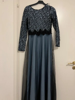 Blå långklänning med spetsdetaljer - Elegant långklänning i mörkblå nyans med långärmad överdel i spets och blommigt mönster. Kjolen är flödande med ett tunt lager svart tyll över blått tyg. Perfekt för dig som vill ha en unik och stilren look.