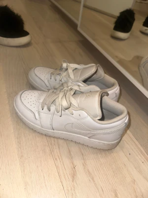 Vita Nike Air Jordan 1 Low sneakers - Säljer ett par vita Nike Air Jordan 1 Low sneakers i skinn med klassisk swoosh på sidan och perforerad tåbox. Skorna har vita snören och en låg silhuett som ger en clean och stilren look. Perfekta för dig som gillar streetwear och sneakers.