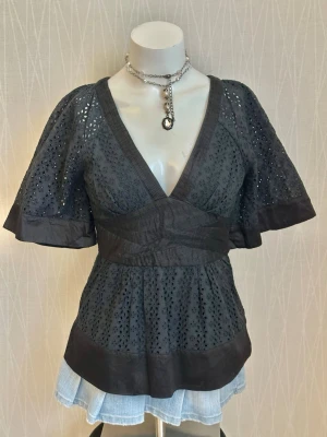 Svart blus med broderade detaljer - Supersnygg svart blus med v-ringning och vida korta ärmar. Blusen har broderade hålmönster och markerad midja som ger en feminin siluett. Perfekt att styla med jeans eller kjol för en trendig look.