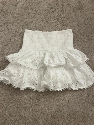 Vit kortkjol med broderad spets - Söt vit kortkjol med broderad spets och volangdetaljer. Kjolen har elastisk midja och är gjord i bomull, vilket gör den både bekväm och luftig. Perfekt för dig som gillar romantiska och feminina plagg.