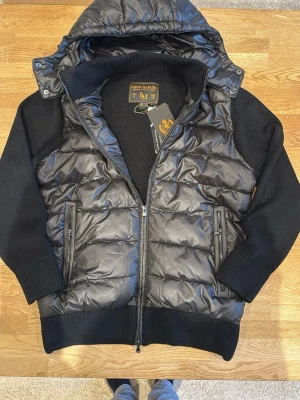 Svart dunväst från Leon Marcel - Ej använd, Snygg svart cardigan från Leon Marcel med stickade ärmar och rygg. Västen har dragkedja framtill, två fickor med dragkedja och en avtagbar huva. Materialmix av dun och stickat ger en modern look. Perfekt för lager-på-lager-stil.