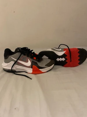 Nike svartvita sneakers med röd detalj - Snygga Nike sneakers i svart, vitt och grått med röda detaljer på sulan. Skorna har en låg profil, klassisk snörning och en slitstark yttersula med mönster för bra grepp. Perfekta för dig som gillar sportig och modern stil.