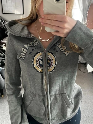 Grå hoodie från Abercrombie & Fitch. LITEN i storleken! - Snygg grå hoodie med dragkedja från Abercrombie & Fitch. Stor broderad logga och emblem på bröstet, samt texten 'Abercrombie New York'. Mjuk bomullsblandning, fickor fram och klassisk huva. Perfekt för en avslappnad och trendig look.
