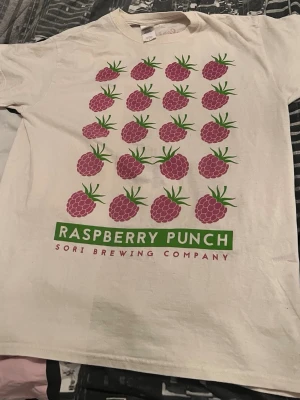 Vit t-shirt Sori Brewing hallontryck - Vit t-shirt från Sori Brewing Company med stort tryck av hallon och texten 'Raspberry Punch' på framsidan. Klassisk passform och rund hals. Perfekt för dig som gillar unika och färgglada prints.