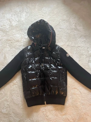 Svart pufferjacka från Moncler - Snygg svart pufferjacka från Moncler med glansig vadderad front och huva med rödvit kant. Jackan har svarta ärmar i mjukare material och dragkedja framtill. Klassisk Moncler-logga på ärmen och justerbar huva med snören.