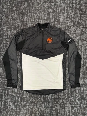 Nike running tröja – Strl S – Ny - Nike Dri-Fit långärmad träningströja i svart och vitt med halv dragkedja. Tröjan har ett orange tryck på bröstet och vit Nike-logga på ärmen. Tillverkad i lätt, tekniskt material som andas och passar perfekt till träning eller att bara ha på sig i vardagen.