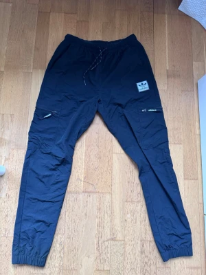 Svarta cargobyxor från Adidas - Svarta cargo byxor från Adidas.  Färg : Matt svart   Byxorna har dragsko vid midjan, reså i midjan och på för anklarna.  Två fickor sidan av låren med dragkedja. En ficka på rumpan med tre sträck (reflex) Byxorna är i ett hårdare prassel material.   Storlek : S , smal passform.  Bredd Midja : 36cm , drar man ut i midjan blir det 44-45cm.  Bredd på höft : 55cm.  Längd på innerben av byxan : 74cm.  Längd på ytterbben : 103cm  Säljer byxorna för att de är för små för mig . Har använt byxorna max 3 