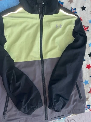 Under Armour vindjacka med huva - Snygg vindjacka från Under Armour med huva, dragkedja och två fickor. Jackan har färgblock i svart, ljusgul och grå samt reflekterande detaljer på axlarna. Tillverkad i lätt och slitstarkt material med meshfoder för extra ventilation.