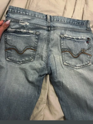 Ljusblå slitna bootcut-jeans Nudie - Säljer ett par ljusblå bootcut-jeans från Nudie med slitningar och trasiga detaljer på både fram- och bakfickor. Jeansen har klassiska fem fickor, orange sömmar och vågigt broderi på bakfickorna. Tillverkade i bomull med en cool vintagekänsla.