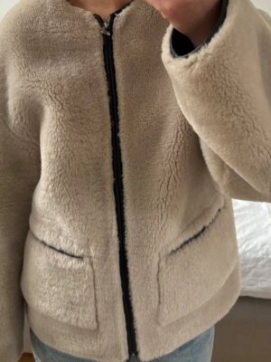Beige teddyjacka med dragkedja - Mysig beige teddyjacka med rak passform och svart dragkedja framtill. Jackan har två stora fickor och mjukt, fluffigt material som ger en avslappnad look. Perfekt för kyliga dagar och enkel att matcha med jeans eller andra basplagg.