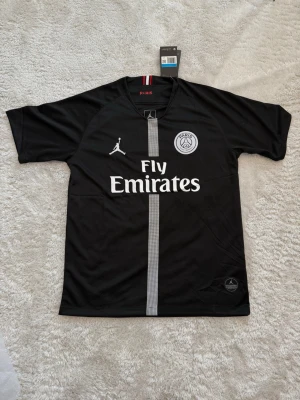 Svart PSG matchtröja Jordan x Nike - Snygg svart PSG matchtröja från samarbetet mellan Jordan och Nike. Tröjan har vita loggor, Fly Emirates-tryck och en grå vertikal rand på framsidan. Tillverkad i lätt och ventilerande material, perfekt för fotboll eller träning.