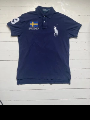 Blå Ralph Lauren Sweden piké - Mörkblå pikétröja från Ralph Lauren med svensk flagga och texten SWEDEN på bröstet och ryggen. Stor vit polospelare på framsidan och siffran 3 på ärmen och ryggen. Klassisk krage och knappar framtill.