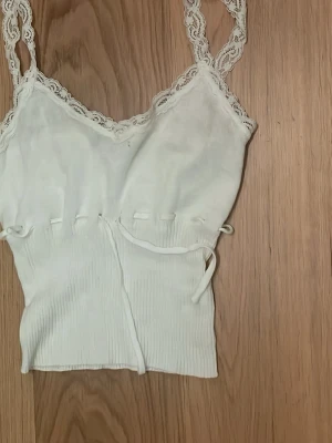 Vit ribbad linnetopp med spets - Söt vit linnetopp med ribbad struktur och spetsdetaljer längs axelbanden och halsringningen. Toppen har snörning i midjan för justerbar passform och är ärmlös. 