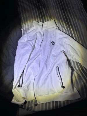 philipp plein zip - Snygg vit zop hoodie från philipp plein med dragkedjor på fickorna och en liten logga på bröstet. Hoodien har hög krage och justerbara snören vid midjan. Perfekt för en clean och sportig look.